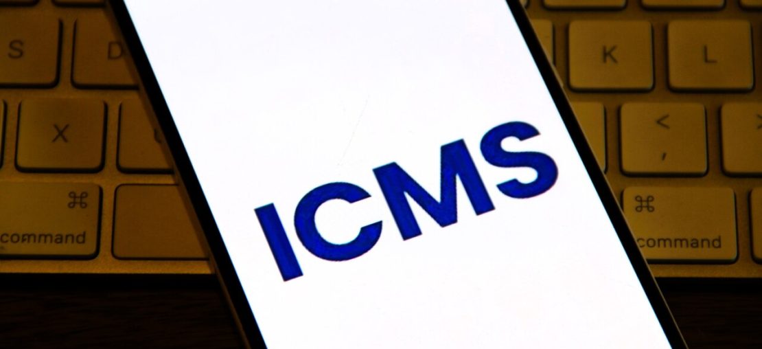 Smartphone exibindo sigla ICMS na tela, representando consulta &agrave; tabela ICMS e c&aacute;lculo de impostos estaduais.