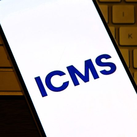 Smartphone exibindo sigla ICMS na tela, representando consulta &agrave; tabela ICMS e c&aacute;lculo de impostos estaduais.