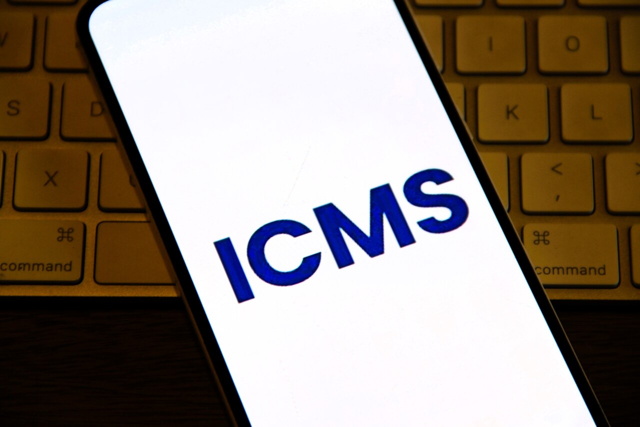 Smartphone exibindo sigla ICMS na tela, representando consulta à tabela ICMS e cálculo de impostos estaduais.